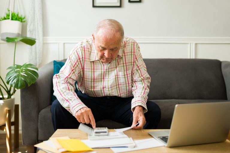 Um idoso faz cálculos com um papel na mão e uma calculadora. Em cima da mesa ha um laptop. Ele tem cabelo, barba e bigode branco, e veste uma camisa cáqui