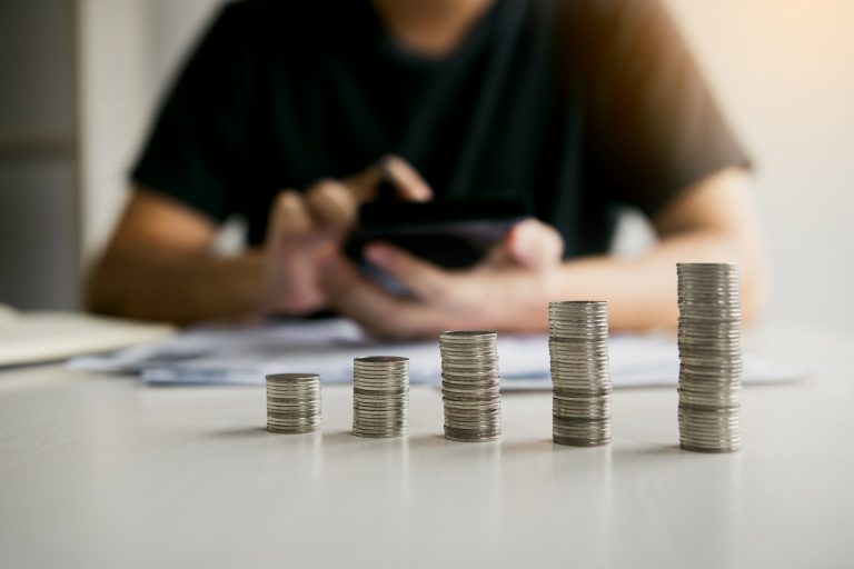 Cinco pequenas pilhas de moedas sobre uma mesa, em destaque na foto, e uma pessoa fazendo contas ao fundo